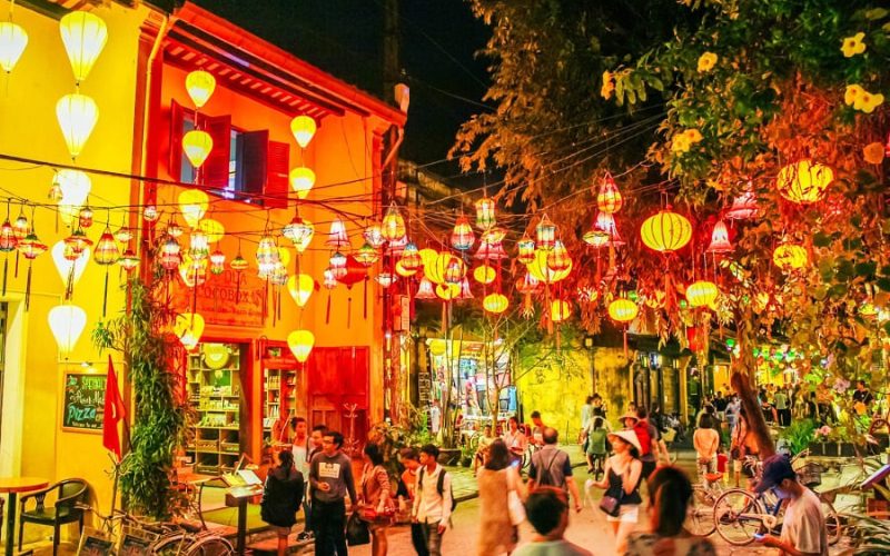 Ruelle animée de Hoi An illuminée par des lanternes colorées suspendues, avec des maisons jaunes typiques et des passants profitant de l’ambiance nocturne festive