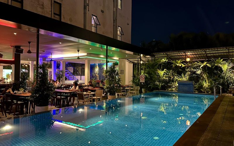 Piscine éclairée de nuit dans un hôtel moderne au Cambodge, entourée de verdure et d’un restaurant en plein air animé sous des lumières colorées