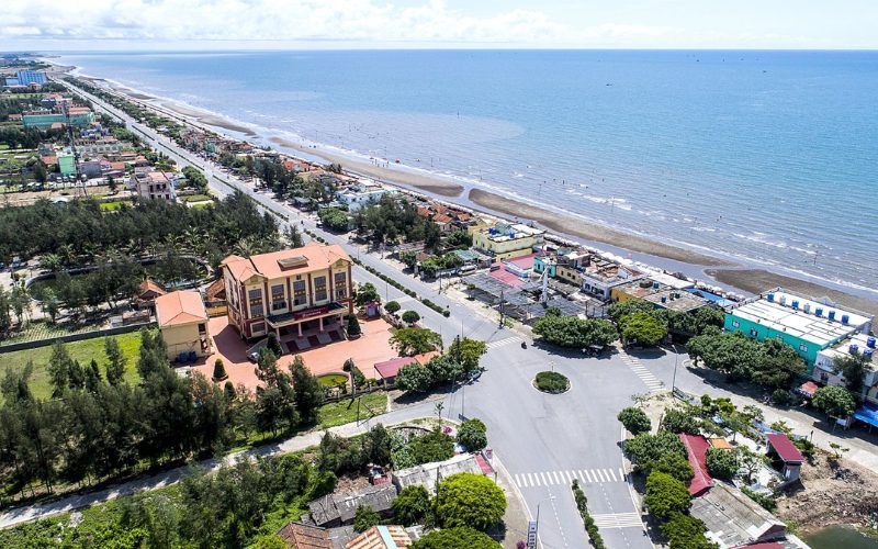 Vue aérienne de la plage de Thinh Long à Nam Dinh, avec ses maisons colorées, sa route côtière bordée d’arbres et la mer calme s’étendant à l’horizon