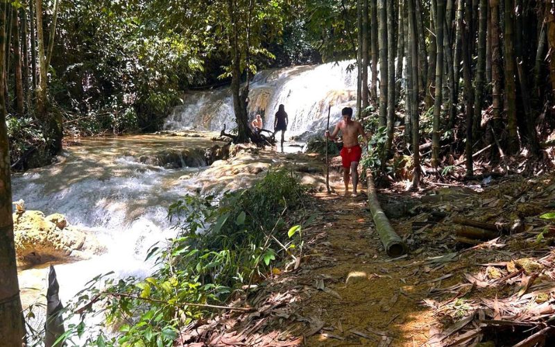 Des visiteurs marchent en maillot de bain le long d’un sentier forestier bordé de bambous, près d’une cascade en terrasses entourée de végétation luxuriante, dans une zone naturelle paisible du Vietnam