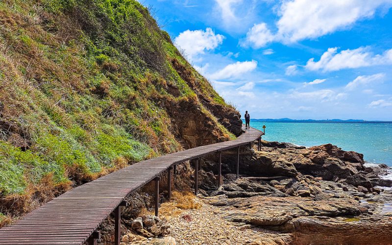 Passerelle en bois longeant une falaise rocheuse au bord de la mer turquoise, sous un ciel bleu éclatant, avec des promeneurs profitant du paysage côtier