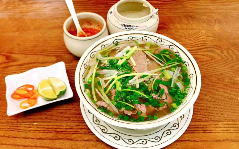 Bol de phở bò de Nam Dinh servi avec des herbes fraîches, du citron vert et des piments tranchés, accompagné de condiments traditionnels vietnamiens