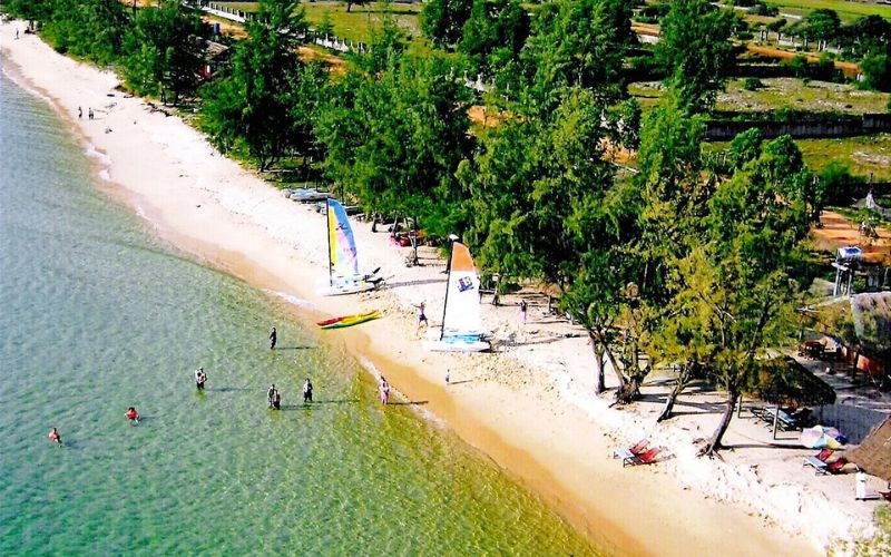 Vue aérienne d’une plage de sable fin bordée d’arbres, où des baigneurs profitent de l’eau claire et de petits voiliers colorés sont amarrés près du rivage