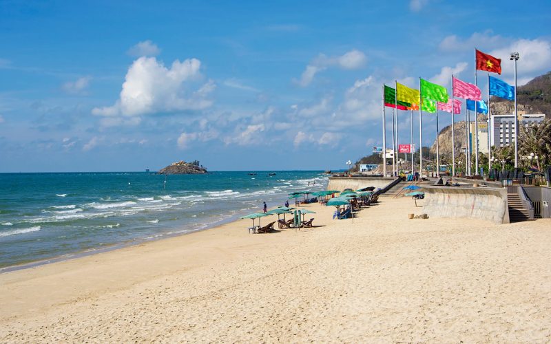 Plage de Back Beach à Vung Tau, avec sable doré, parasols alignés le long du rivage, mer turquoise sous un ciel bleu et rangée de drapeaux colorés flottant au vent