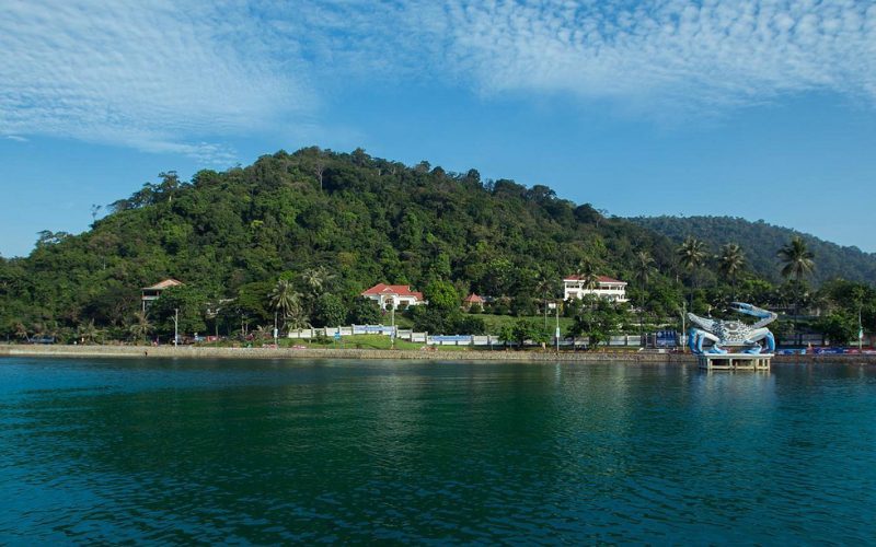 Vue panoramique de la côte de Kep, avec sa célèbre statue de crabe géant au bord de l’eau, des collines verdoyantes en arrière-plan et un ciel bleu parsemé de nuages