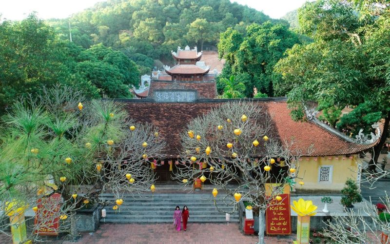 Vue aérienne de la pagode Con Son à Hai Phong, avec son toit traditionnel en tuiles rouges entouré de collines verdoyantes. À l’entrée, des arbres décorés de lanternes jaunes et deux femmes en áo dài vietnamien ajoutent une touche culturelle et colorée à la scène