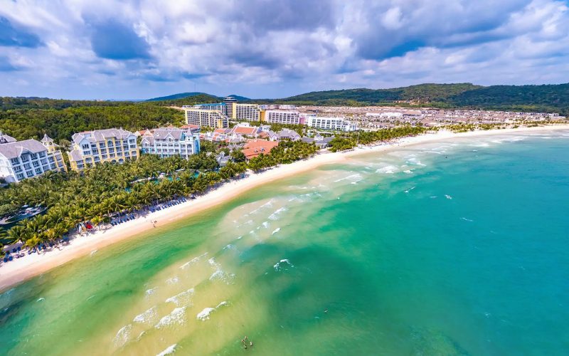 Vue aérienne spectaculaire de la plage de sable blanc de Phu Quoc bordée d’eaux turquoise, avec des complexes hôteliers de luxe nichés entre les palmiers et la forêt tropicale en arrière-plan