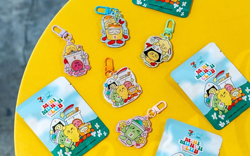 Porte-clés colorés et sachets d’emballage de la collection “Mini Munch Club” de 7-Eleven, avec des personnages mignons inspirés de snacks, posés sur une table jaune vive