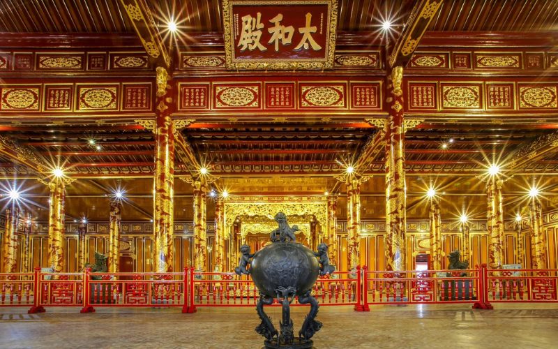 Intérieur somptueux du palais Thai Hoa dans la Cité impériale de Hué, orné de colonnes rouges et dorées sculptées de motifs de dragons, avec un trône impérial au fond et une grande plaque calligraphiée suspendue au plafond
