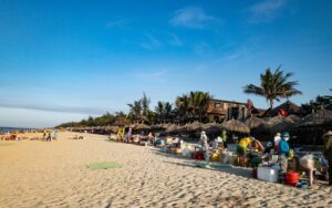 Plage animée avec des vendeurs installés sous des parasols en chaume, des cabanes en bois, des palmiers et de nombreux vacanciers profitant du soleil en fin de journée