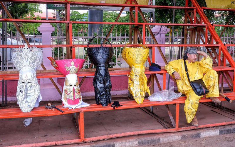 Masques traditionnels colorés du festival Phi Ta Khon alignés sur un banc en métal, à côté d’un participant en costume jaune se reposant dans les gradins