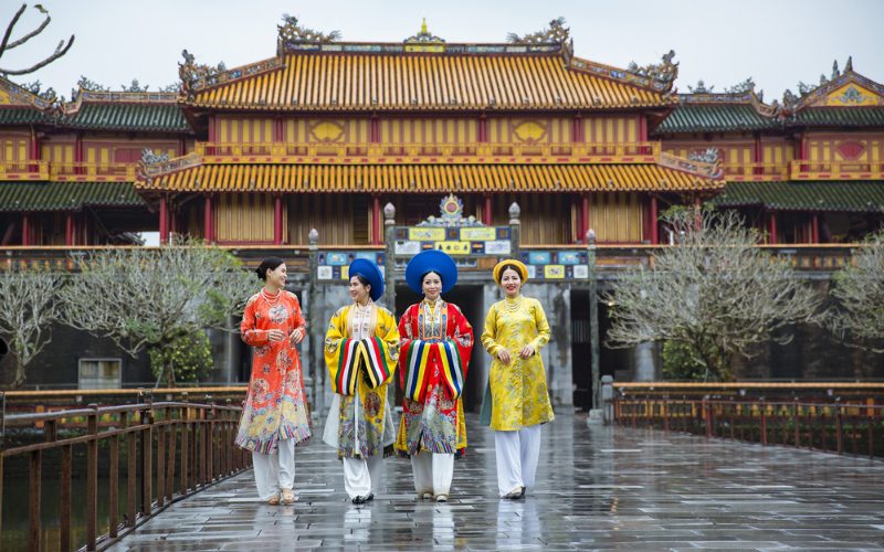 Quatre femmes en áo dài traditionnel vietnamien coloré marchant sur un pont devant la porte Ngo Mon de la Cité impériale de Hué, sous un ciel gris