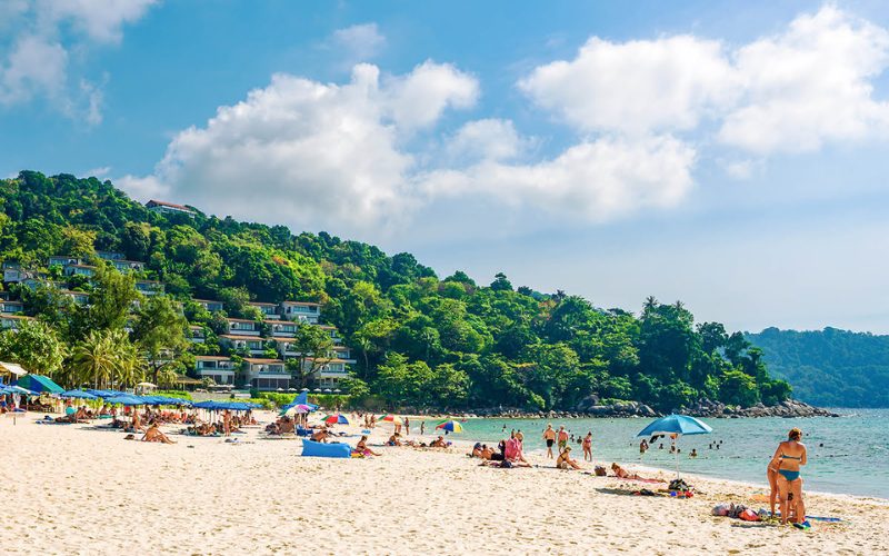 Plage animée de sable blanc à Phuket, Thaïlande, avec des vacanciers se relaxant sous des parasols et nageant dans la mer turquoise, entourée de collines verdoyantes