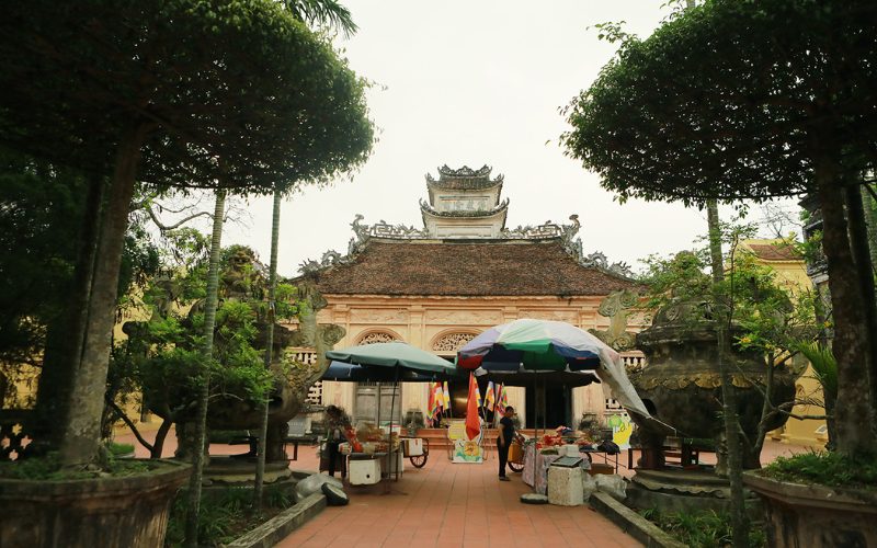 Temple ancien au toit de tuiles rouges à Nam Dinh, entouré de grands arbres taillés et d’encensoirs en pierre, avec des étals colorés et des drapeaux bouddhiques à l’entrée
