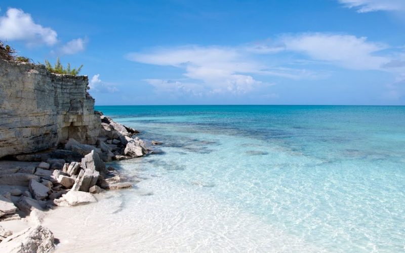 Plage de sable blanc bordée de rochers avec une mer turquoise et limpide sous un ciel bleu ensoleillé, offrant un paysage côtier paisible et préservé