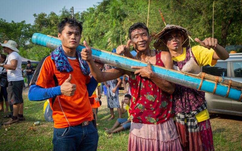 Participants souriants et vêtus de manière festive portant une fusée artisanale en bambou lors du festival des fusées Bun Bang Fai au Laos