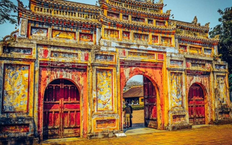 Porte ancienne aux couleurs rouge et ocre de la Cité impériale de Hué, ornée de motifs traditionnels vietnamiens et de fresques de dragons, avec deux visiteurs entrant par l’arche centrale