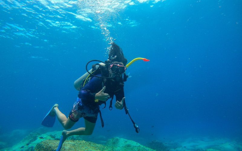 Plongeur en combinaison avec masque et bouteille d’oxygène faisant un signe de main sous l’eau