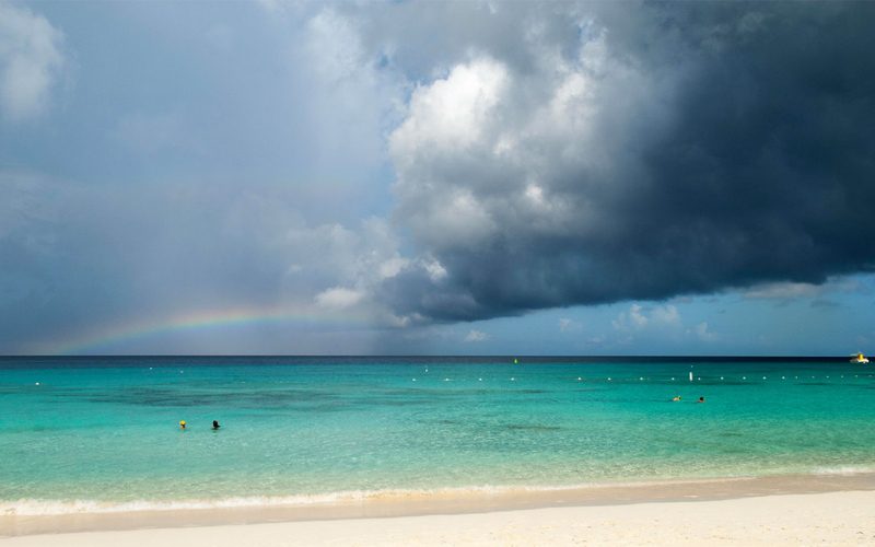 Plage de sable blanc avec une mer turquoise sous un ciel orageux, un arc-en-ciel discret apparaissant à l’horizon et quelques baigneurs profitant de l’ea