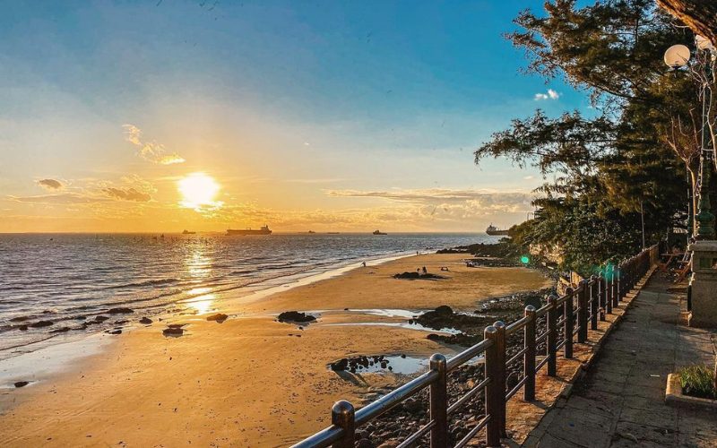Coucher de soleil sur la plage de Bai Dau à Vung Tau, avec sable doré, mer calme, silhouettes de navires au loin et promenade bordée d’arbres et de rambardes.