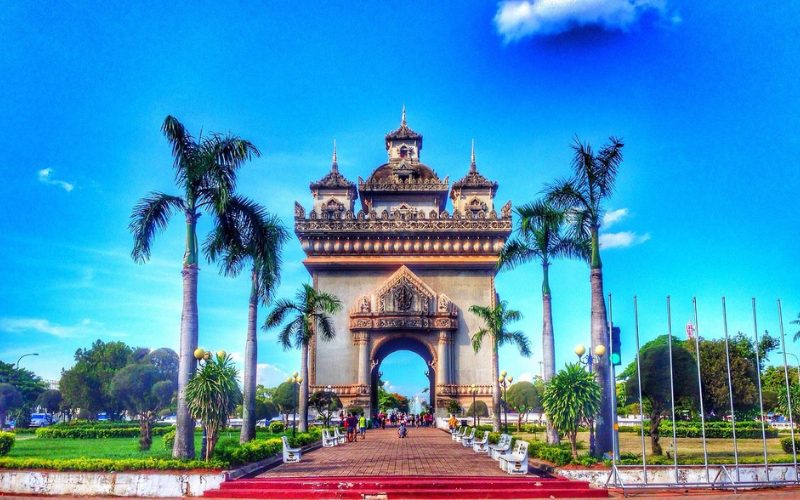 Vue frontale du Patuxai, l'Arc de Triomphe de Vientiane au Laos, entouré de palmiers et d’un jardin public, sous un ciel bleu éclatant