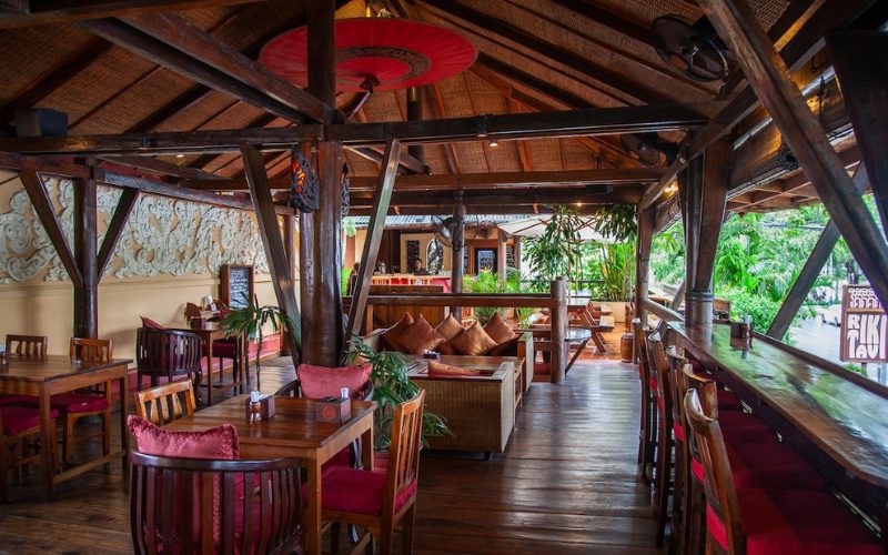 Intérieur chaleureux du restaurant Rikitikitavi à Kampot, avec une décoration en bois traditionnel, des tables et chaises en bois foncé, des coussins colorés