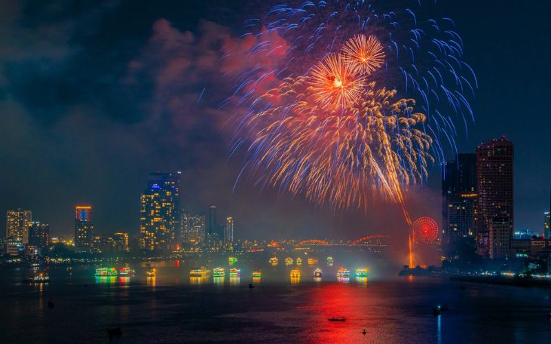 Feux d’artifice éclatants illuminant le ciel nocturne de Da Nang au-dessus de la rivière Hàn, avec en arrière-plan les gratte-ciel lumineux, la grande roue rouge et une flottille de bateaux décorés naviguant sur l’eau scintillante