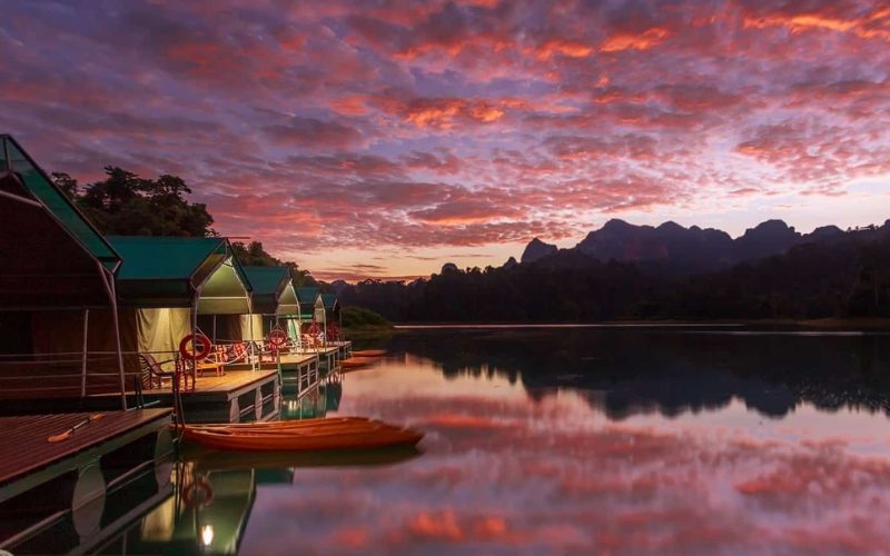 Bungalows flottants au bord d’un lac calme sous un ciel aux couleurs flamboyantes du coucher de soleil, entourés de montagnes et de forêt tropicale