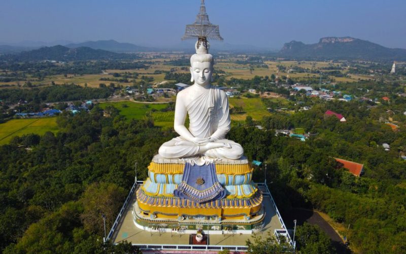 Grande statue blanche de Bouddha en position de méditation, trônant au sommet d’un temple coloré au milieu d’un paysage rural verdoyant en Thaïlande