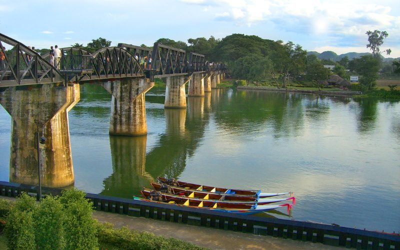 Vue du célèbre pont de la rivière Kwai à Kanchanaburi, en Thaïlande, avec des visiteurs marchant dessus et des bateaux colorés amarrés sur l’eau calme en contrebas