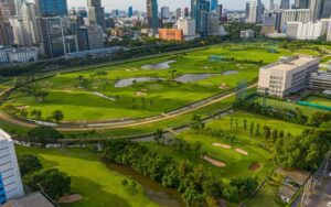 Vue aérienne d’un parcours de golf en plein centre-ville de Bangkok, entouré de gratte-ciel modernes, de bâtiments commerciaux et de verdure luxuriante, offrant un contraste unique entre nature et urbanisme