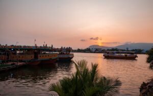 Bateaux de croisière illuminés flottant sur la rivière de Kampot au coucher du soleil, avec en arrière-plan les montagnes et le ciel teinté d’orange