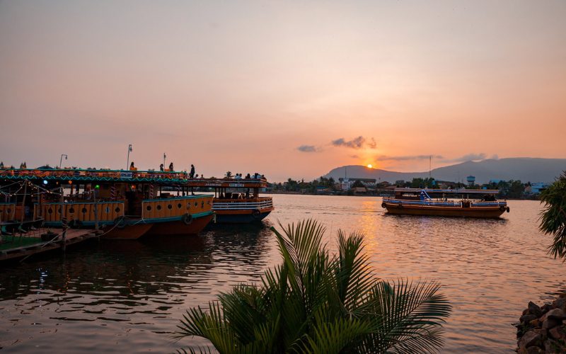 Bateaux de croisière illuminés flottant sur la rivière de Kampot au coucher du soleil, avec en arrière-plan les montagnes et le ciel teinté d’orange