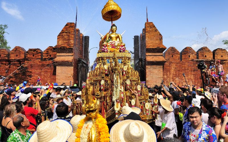 Procession de Songkran à Chiang Mai avec une statue dorée de Bouddha richement décorée portée devant la porte Tha Phae, entourée d’une foule de participants aspergeant d’eau dans une ambiance festive et traditionnelle