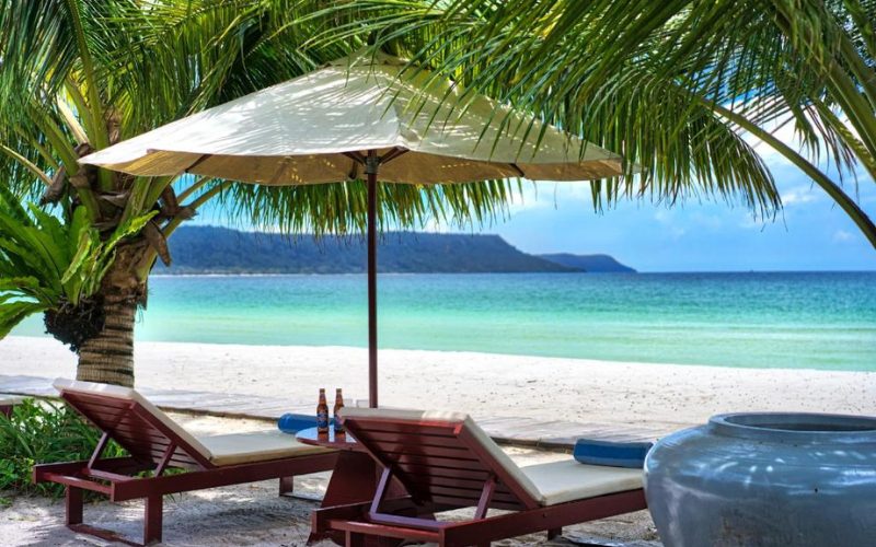 Deux transats en bois sous un parasol blanc, installés à l’ombre des palmiers sur une plage de sable fin face à la mer turquoise à Koh Rong