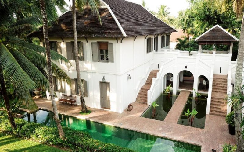 Vue extérieure d’un hôtel de charme à Luang Prabang, installé dans une maison coloniale blanche entourée de palmiers, avec piscine verte et allées en briques rouges, dans un jardin luxuriant