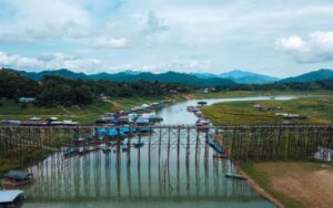 Vue aérienne du pont en bois Mon Bridge à Sangkhlaburi, traversant une rivière paisible entourée de maisons flottantes et de collines verdoyantes