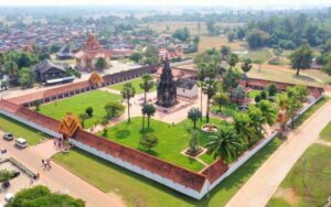 Vue aérienne d’un temple historique au Laos, entouré de murs blancs, de pelouses verdoyantes, de palmiers et de bâtiments traditionnels, au cœur d’un paysage rural paisible