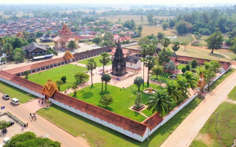 Vue aérienne d’un temple historique au Laos, entouré de murs blancs, de pelouses verdoyantes, de palmiers et de bâtiments traditionnels, au cœur d’un paysage rural paisible