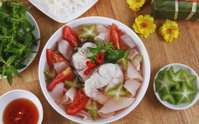 Bol de soupe de poisson Bagridae de Vinh Long, garni de tomates, caramboles et légumes frais, accompagné de vermicelles de riz, herbes aromatiques et sauce nuoc-mam