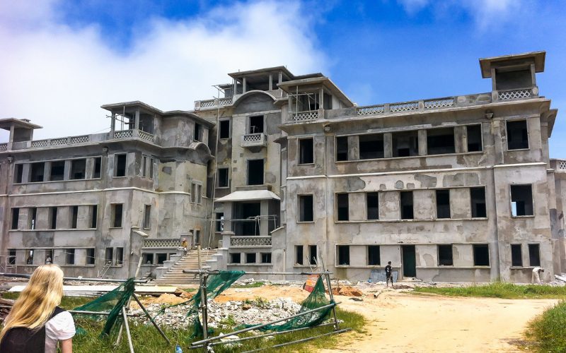 Façade du vieux casino abandonné de la station de Bokor Hill, à Kampot, Cambodge, avec ses murs défraîchis et ses fenêtres sans vitres, entouré de gravats et de végétation, sous un ciel bleu partiellement nuageux