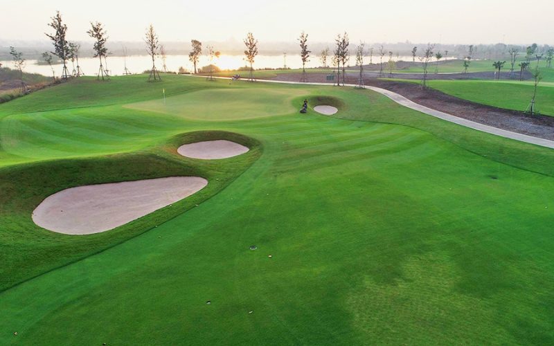Vue d’un green de golf au lever du soleil, avec plusieurs bunkers de sable bien dessinés, des fairways parfaitement entretenus et un lac scintillant en arrière-plan