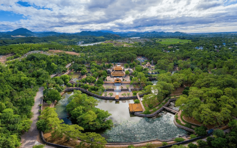 Vue aérienne du tombeau de l’empereur Tu Duc à Hué, Vietnam, entouré de forêts verdoyantes, de jardins paisibles et d’un étang en forme de lotus sous un ciel partiellement nuageux.