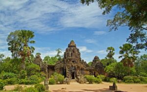 Temple ancien en pierre entouré de végétation tropicale luxuriante, sous un ciel bleu parsemé de nuages, dans un cadre paisible au Cambodge