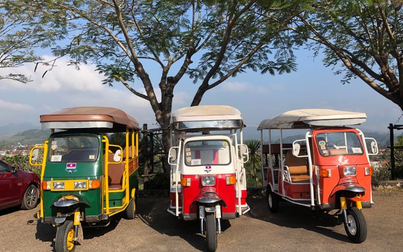 Trois tuk-tuks colorés stationnés côte à côte sous des arbres, prêts à transporter les visiteurs pour explorer la ville
