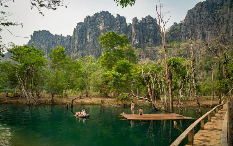 Baignade dans une rivière naturelle entourée de falaises karstiques et de forêt, avec des visiteurs se relaxant sur un radeau et une passerelle en bois longeant l’eau