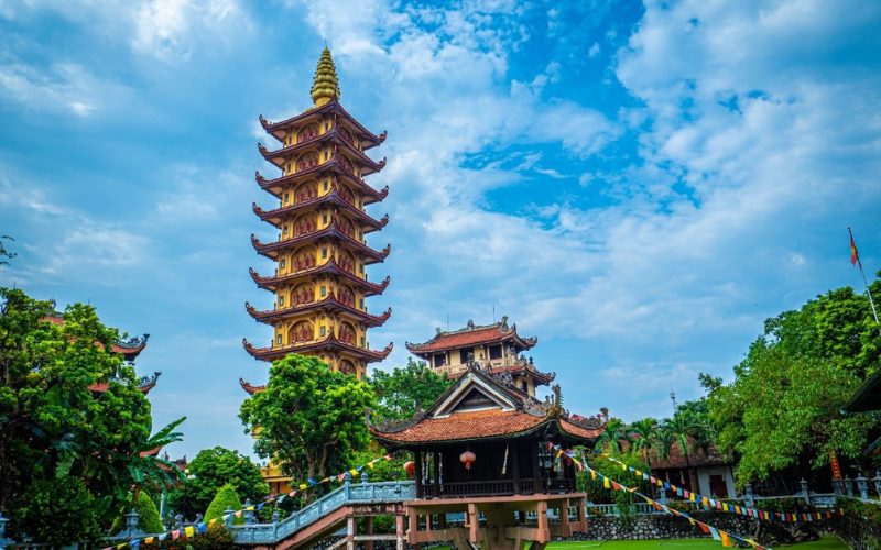 Pagode à Hai Phong avec une tour à plusieurs étages de style bouddhiste s’élevant vers le ciel bleu. Au premier plan, un petit temple sur pilotis au-dessus d’un bassin vert, décoré de lanternes et de fanions colorés, entouré de jardins et d’escaliers ornés