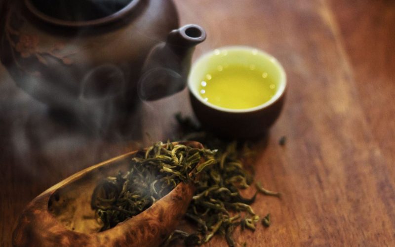 Tasse de thé vert fumant servie à côté d’une théière en terre cuite et de feuilles de thé séchées, posées sur une table en bois, évoquant la tradition du thé vietnamien