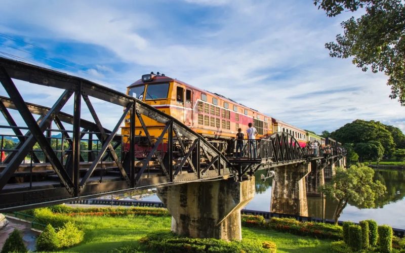 Train jaune et rouge traversant le célèbre pont de la rivière Kwaï à Kanchanaburi, entouré de verdure et reflété dans l’eau sous un ciel bleu