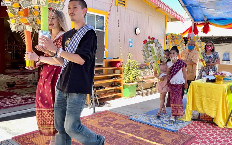 Participants en tenue traditionnelle laotienne portant des arbres à offrandes lors d’une cérémonie religieuse en plein air, dans une ambiance festive et colorée